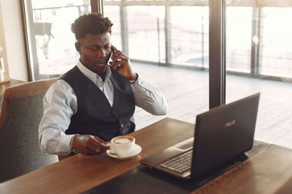 Man with a coffee, laptop, and cellphone sitting in a cafe next to a window